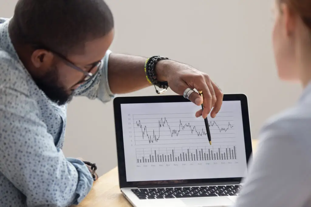 A business professional points to a line and bar graph on a laptop screen while discussing performance data with a colleague, representing operations analysis and decision-making.